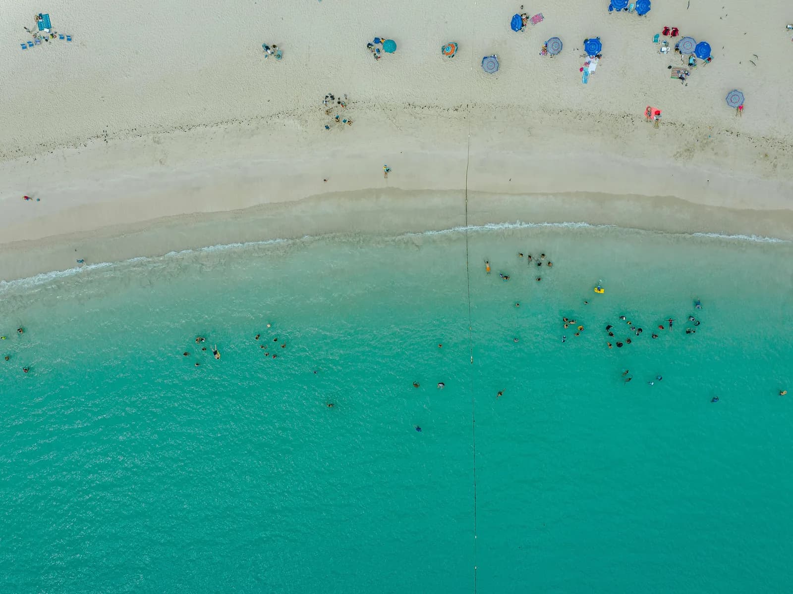 An aerial view of swimmers and umbrellas on a white beach beside turquoise water