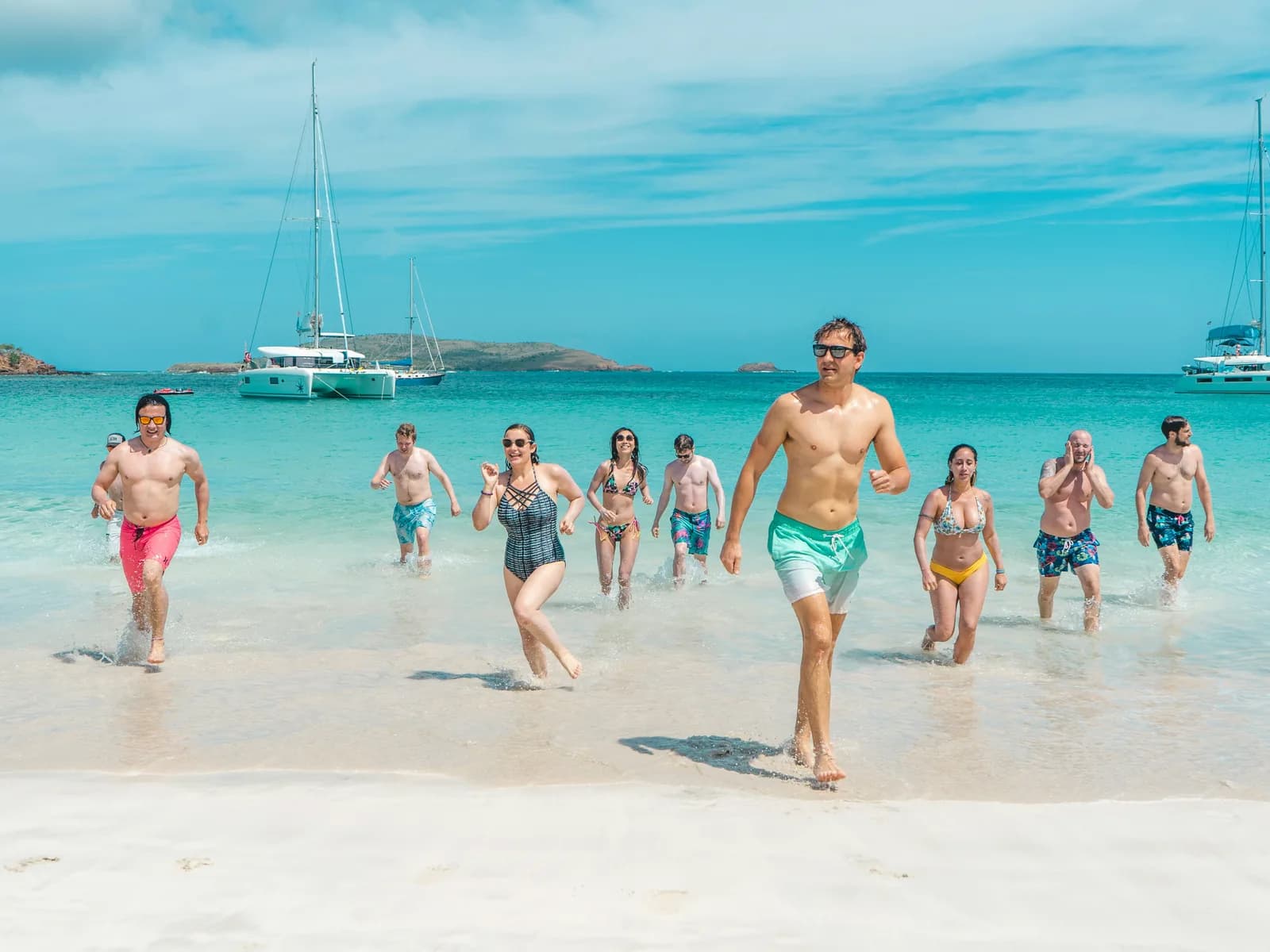 A group running out of the sea together on a beach with catamarans anchored behind